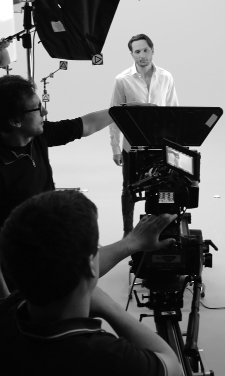 Photo en noir & blanc d'une caméra en train de filmer un comédien avec l'équipe technique